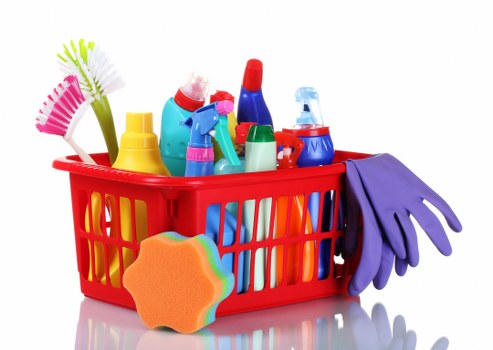 A red plastic shopping basket filled with various cleaning and household items, including several spray bottles in different colours (blue, red, green), a yellow bottle, brushes with pink, green, and white bristles, and a purple pair of rubber gloves draped over the side of the basket. In front of the basket, there is an orange cleaning sponge with a wave-shaped rainbow-colored edge. The items are placed on a clean, reflective surface with a plain white background, indicative of a typical domestic or storage environment for disposal or collection by a rubbish removal service such as Commercial Waste Removal Barnet, serving the Barnet area in North London.