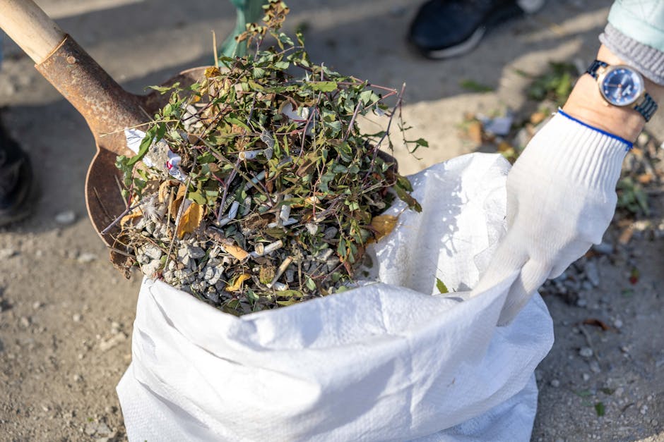 Two large black plastic rubbish bags filled with waste are placed side by side on the pavement next to a curb, with one bag slightly leaning against a black metal fence and the other resting flat. The