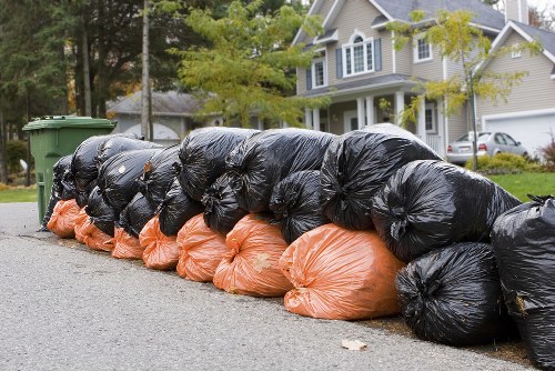A row of tightly packed rubbish bags lined up along the edge of a residential street, with black bags on top and orange bags underneath, on a paved asphalt surface. In the background, a green wheelie bin is visible, along with a driveway leading to a modern, light-colored house with a gabled roof, situated in a suburban neighbourhood with trees and parked cars. The scene suggests rubbish collection or disposal, relevant to commercial waste removal services in Barnet, with the bags likely containing household or business waste awaiting removal or collection. The environment appears clean and orderly, and the outdoor setting is captured during daylight with natural lighting.